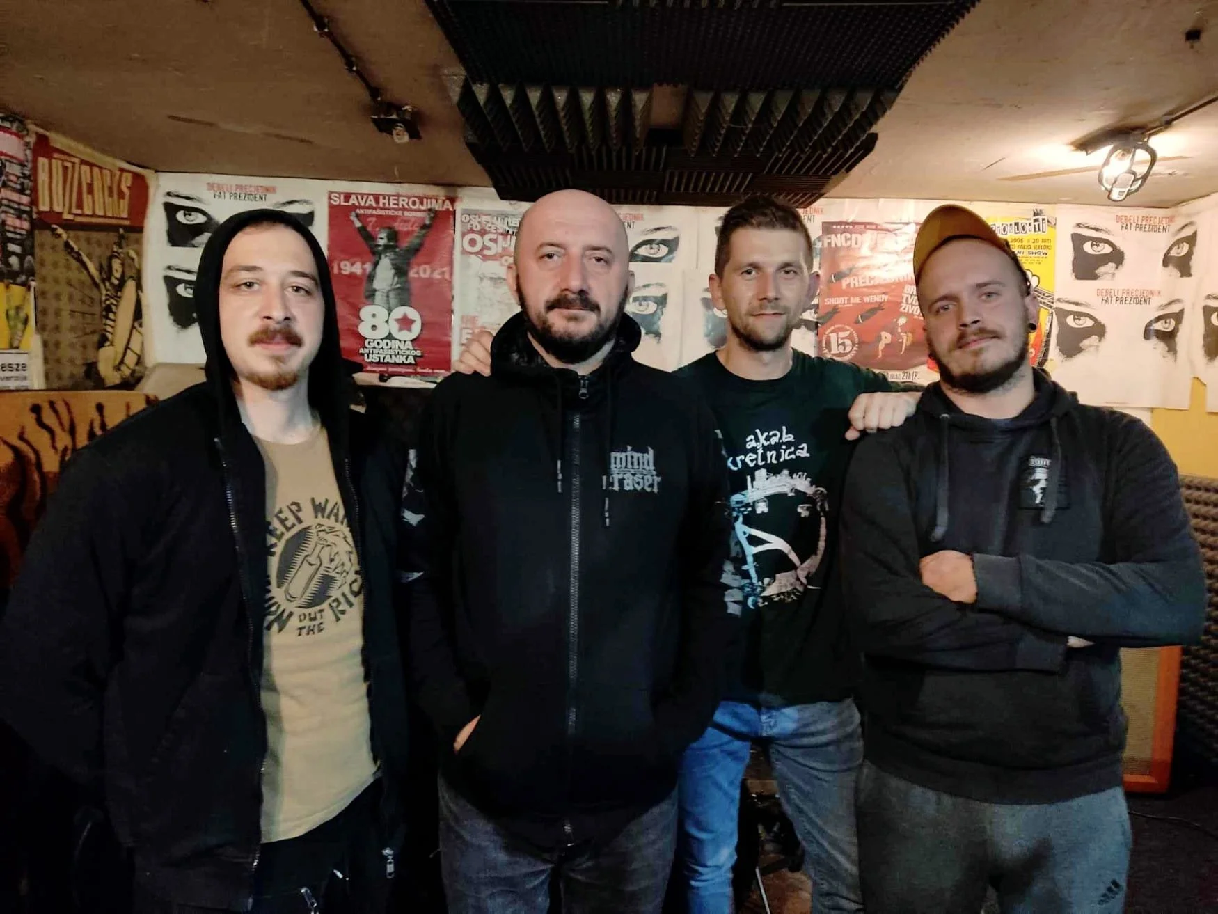 Four men standing together in a dimly lit room with posters on the walls, posing for a photo. They are dressed casually, some with hoodies and jackets.