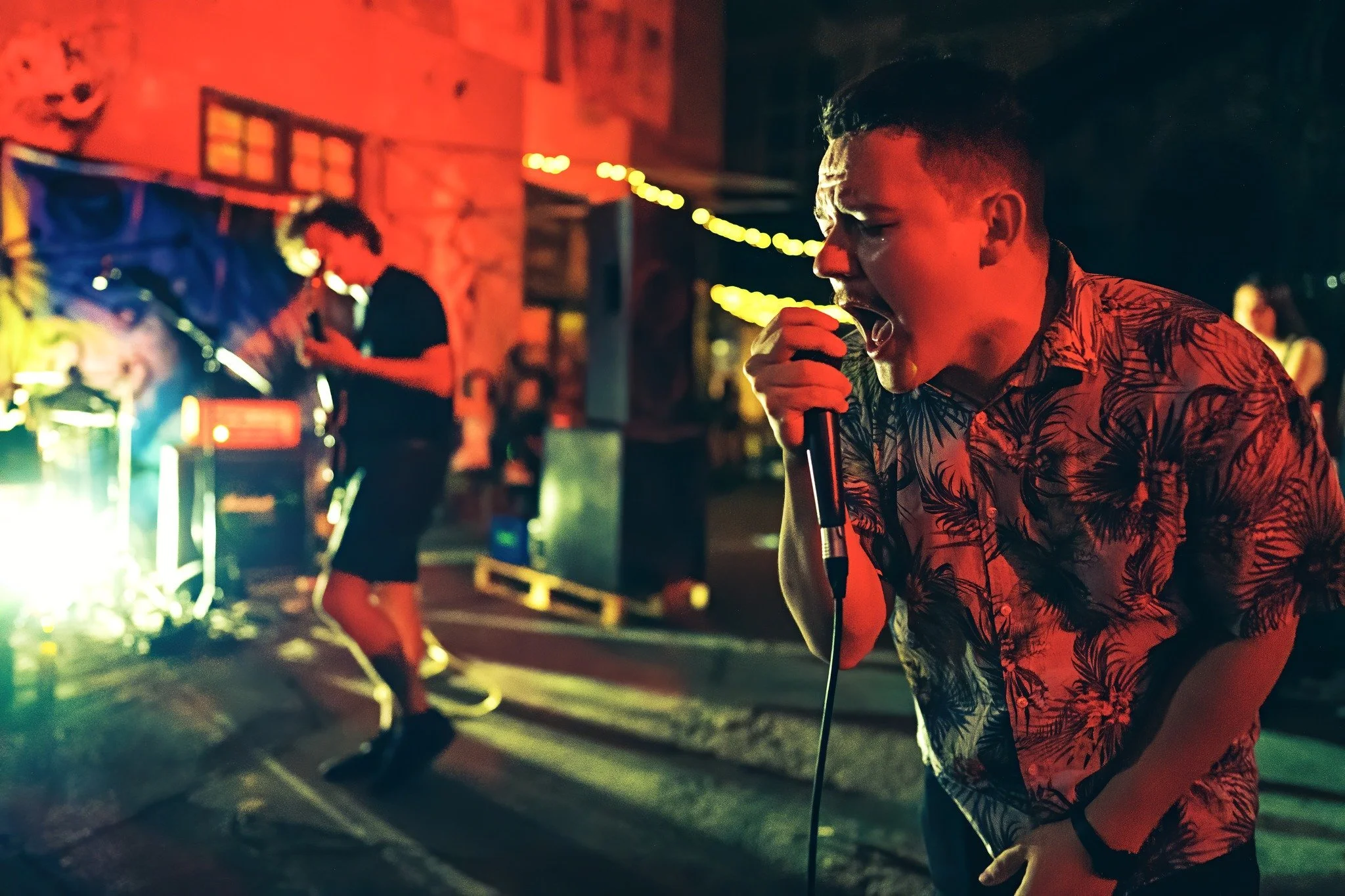 Young man singing into a microphone at a nighttime outdoor event with colorful lighting, other performers and a brick building in the background.