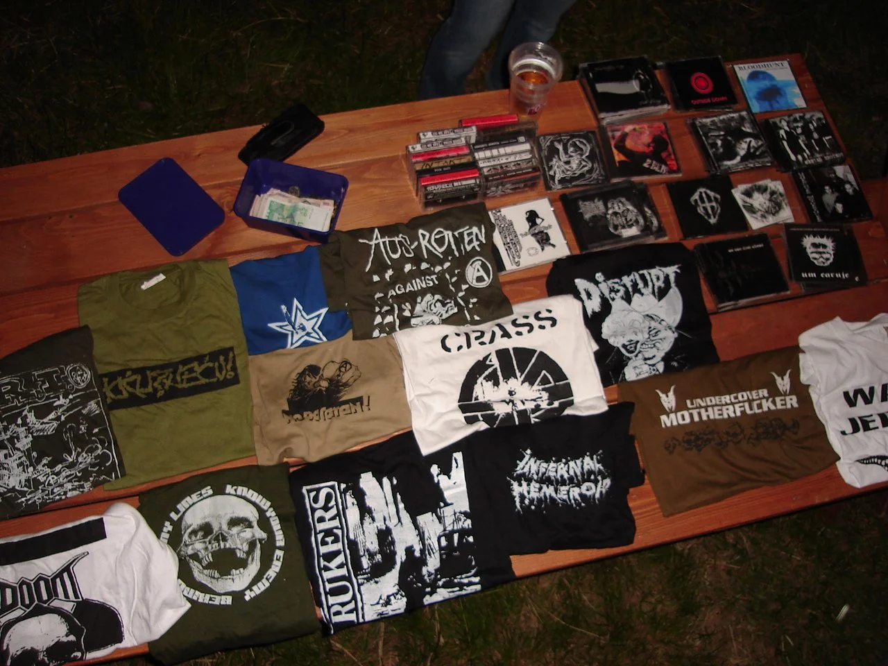 Table with band T-shirts, CDs, and a drink on it, outdoors at night.