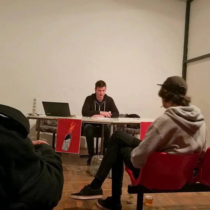 A young man sits at a table with a laptop, participating in a discussion or presentation. There are two other individuals seated nearby, listening, in a room with a plain background. Posters with a flame and bottle image are on the front of the table