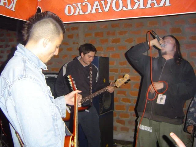 Three musicians performing outdoors in front of a brick wall and a red banner with white text. One is singing into a microphone, and two are playing guitars.
