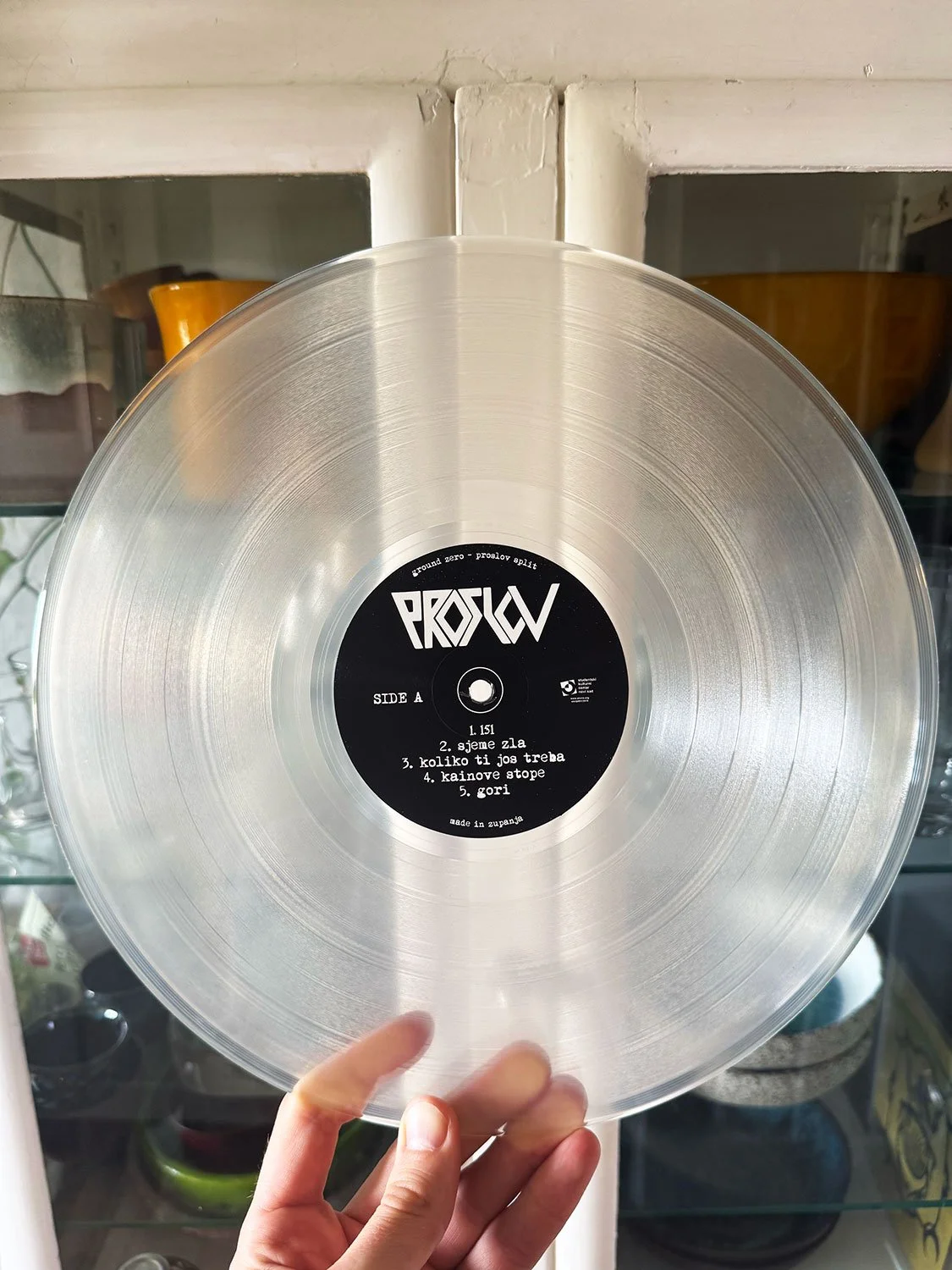 A transparent vinyl record with a black label in the center, held by a person's hand against a background of shelves with various kitchen items.