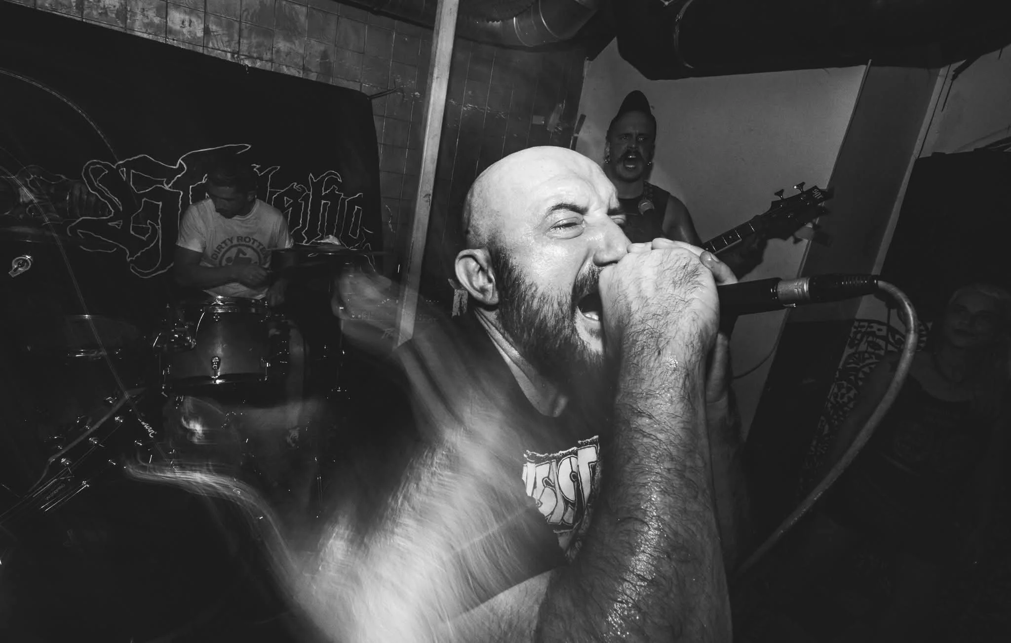 Black and white photo of a band performing live, with a vocalist singing into a microphone, a guitarist in the background, and a drummer behind a drum kit.