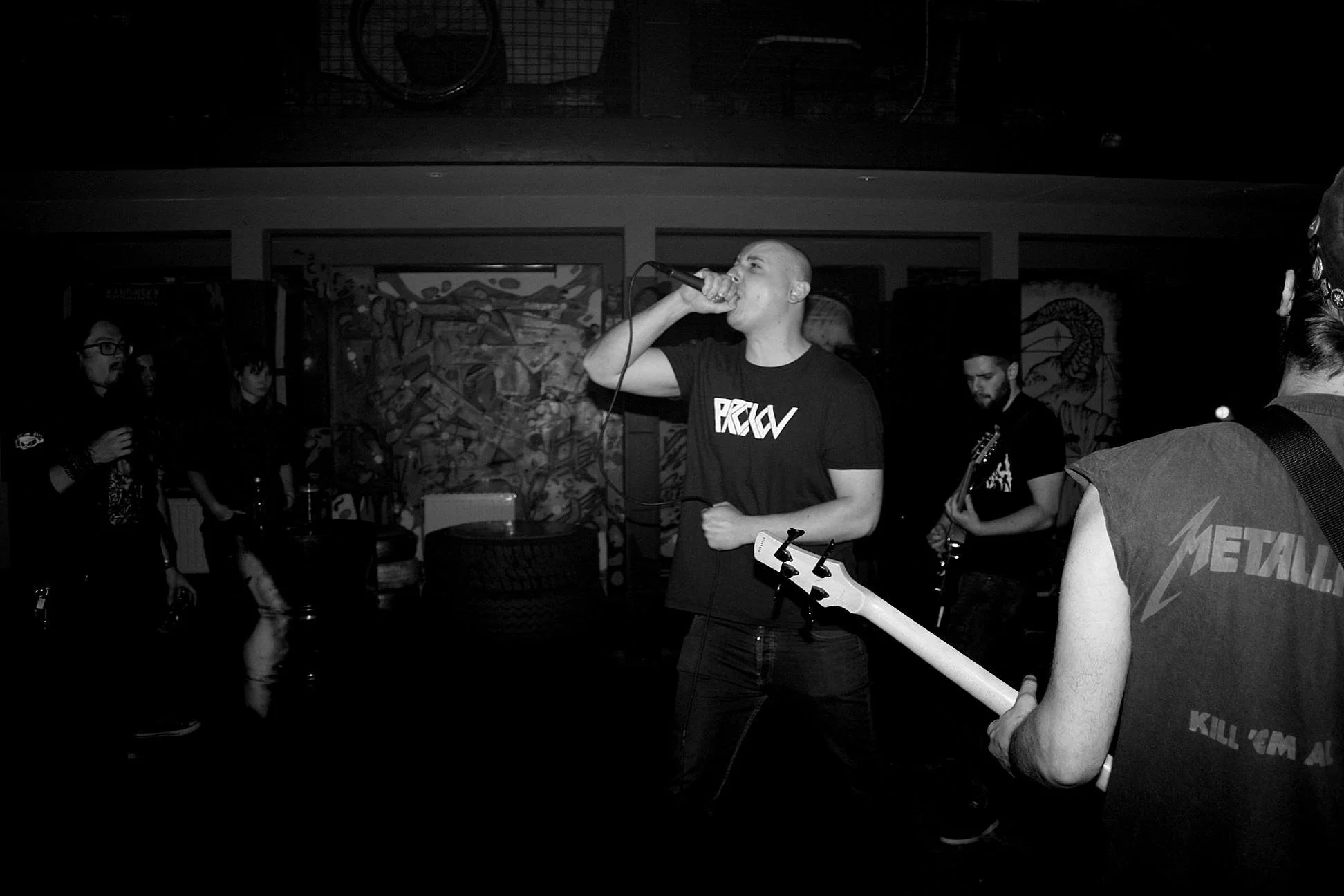 A black and white photo of a live music performance in a small venue. A young man is singing into a microphone, wearing a dark t-shirt, with a bass guitar in front of him. Several other band members and audience members are visible in the background,