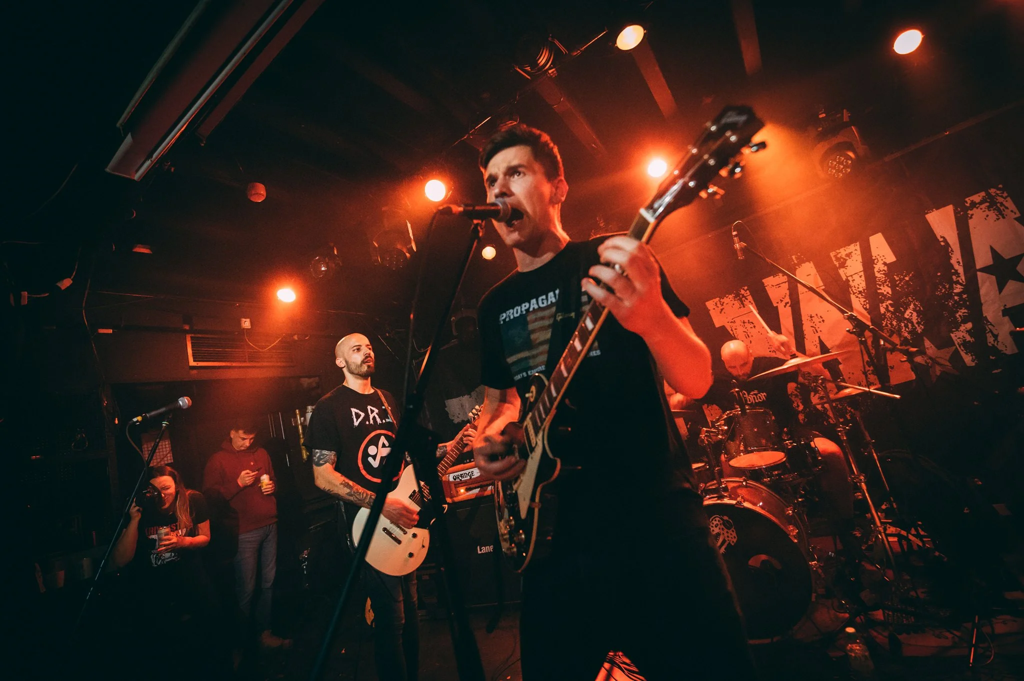 A band performs live on stage in a dimly lit venue with red and orange lighting. The lead singer, holding a microphone in one hand and playing an electric guitar with the other, appears to be singing passionately. Other band members are visible in th