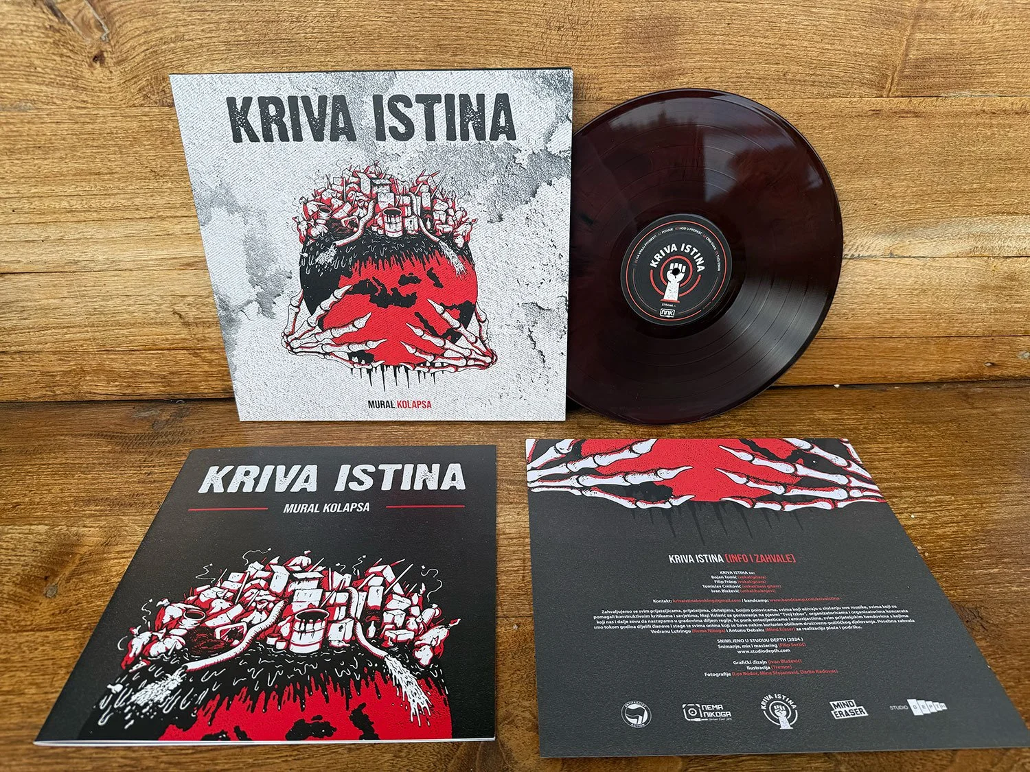 A vinyl record, two flyers, and a large album cover with artwork featuring a red heart with multiple skeletal hands around it and containing numerous buildings on top, laid out on a wooden surface.