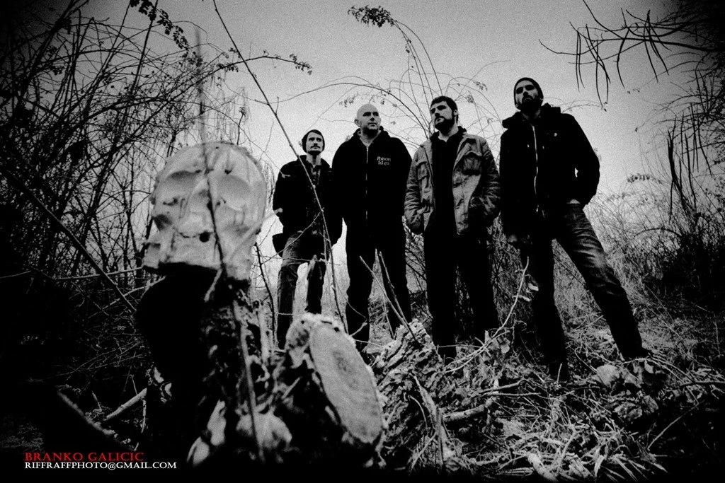 Four men standing in a wooded, dark outdoor area with overgrown plants and a tree with a skull in the foreground, all dressed in dark clothing, giving a moody, mysterious vibe.