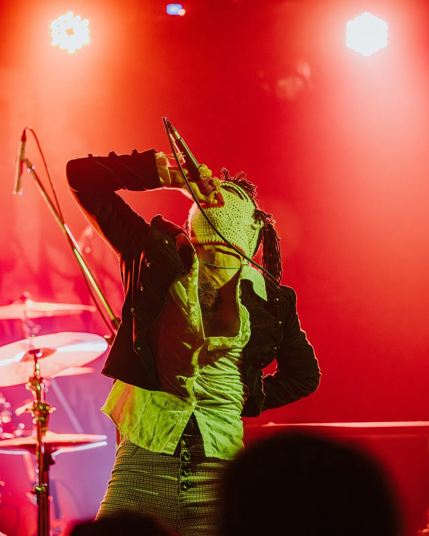 A performer singing or speaking into a microphone on stage, wearing a yellow knit hat with dreadlocks, a black jacket, a yellow scarf, and high-waisted checkered pants, with dramatic stage lighting.