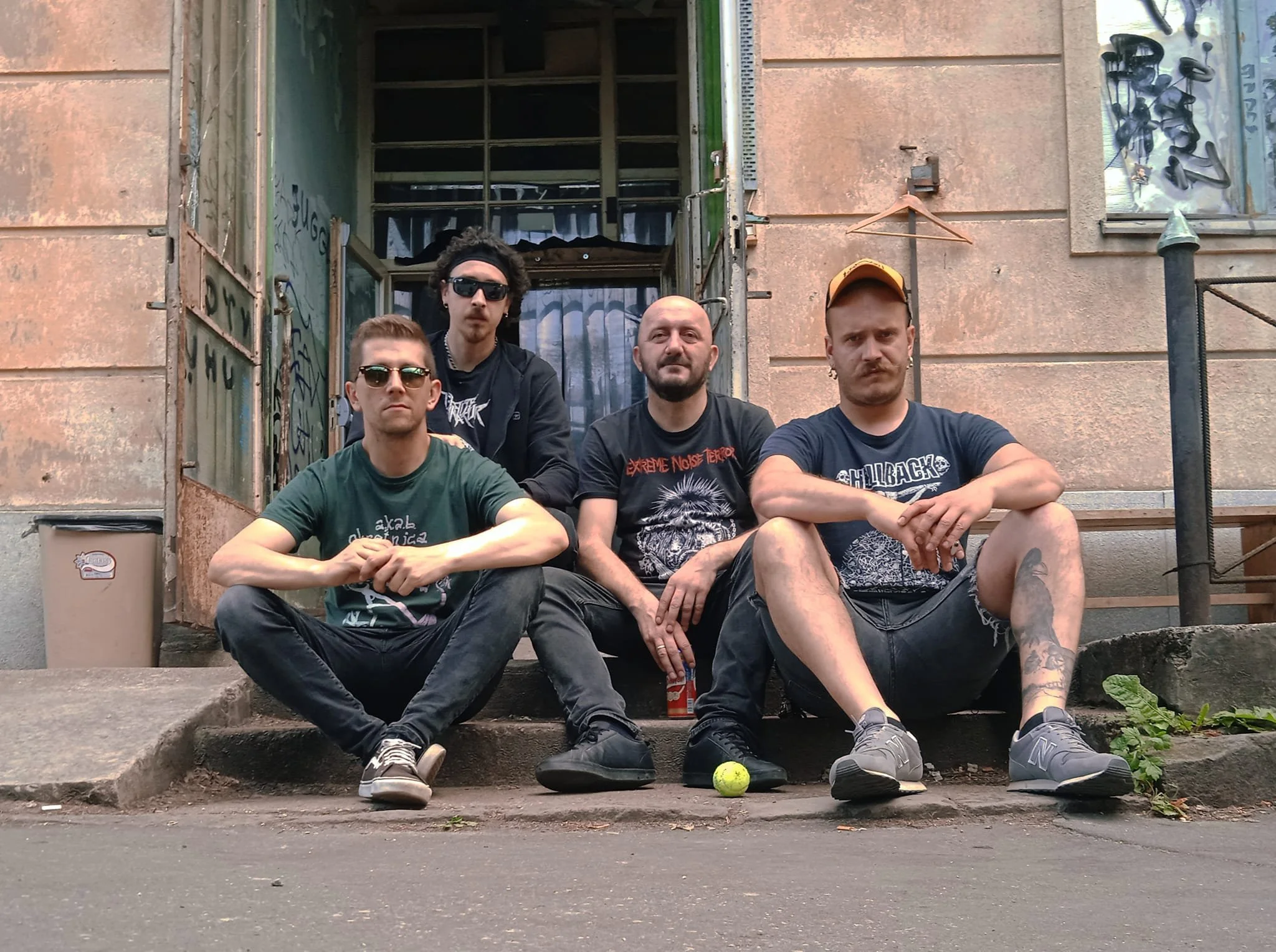 Four young men sitting on concrete steps outside a building with a weathered facade. Two of the men are wearing sunglasses, and they are dressed in casual clothing, including graphic T-shirts, shorts, and sneakers. One man has a tattoo on his leg, and another is wearing a baseball cap. There is a small tennis ball in front of them on the ground. The building behind them has a window covered with film or plastic, and some graffiti is visible on the wall. The photo has an urban, gritty vibe.