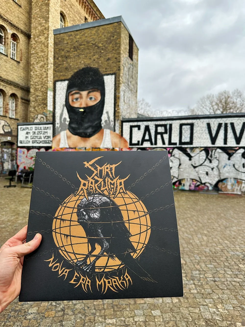 A person holds a black album cover in the foreground featuring a graphic of a black bird with a lion's head, the band's name 'Surfacing' and the phrase 'Nova Era Mraka'.