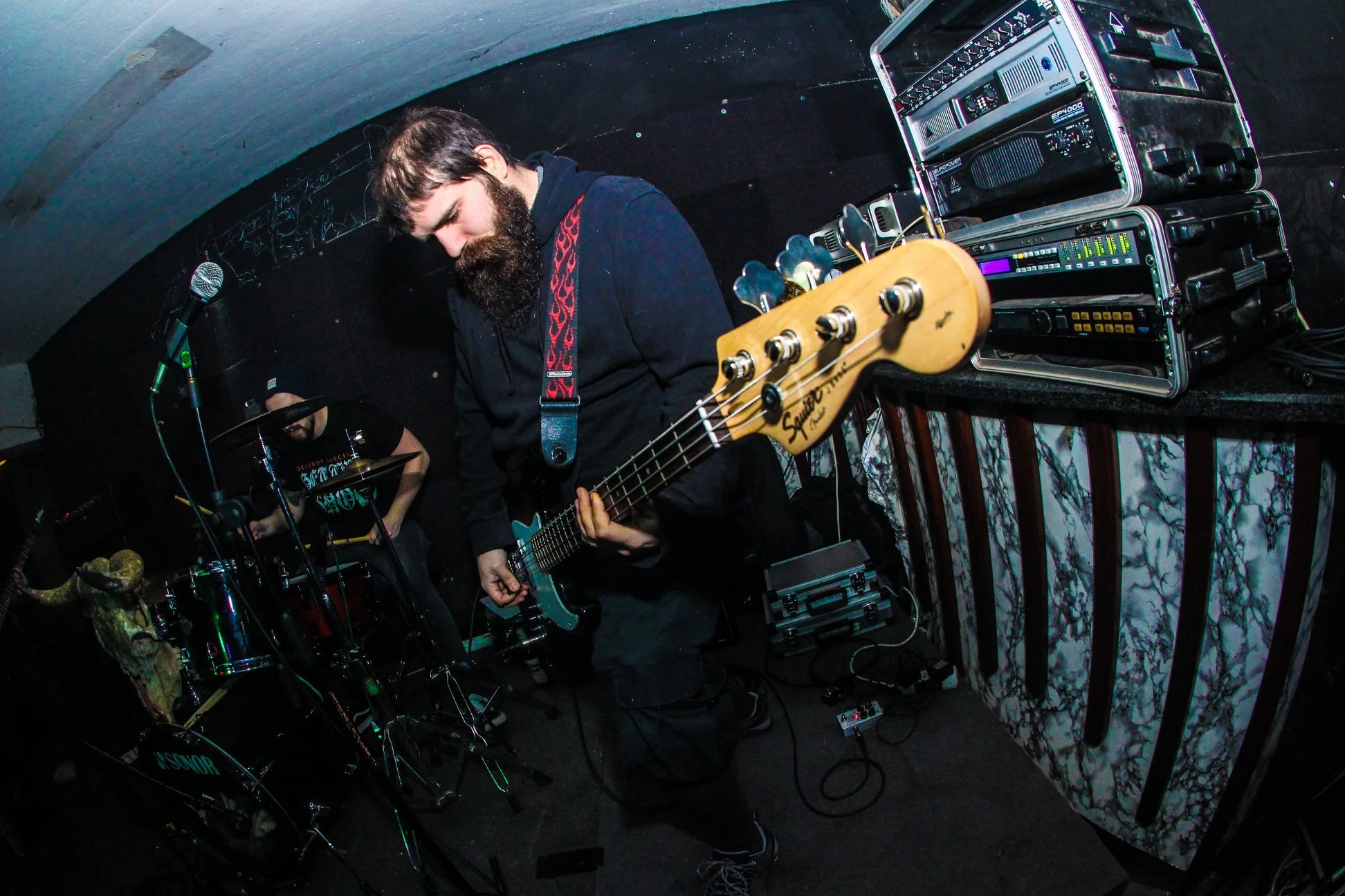 A musician with a beard playing an electric guitar on stage, with a drum set and audio equipment in the background.