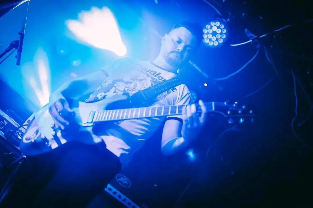 A male guitarist playing an electric guitar on stage with blue lighting and stage equipment in the background.