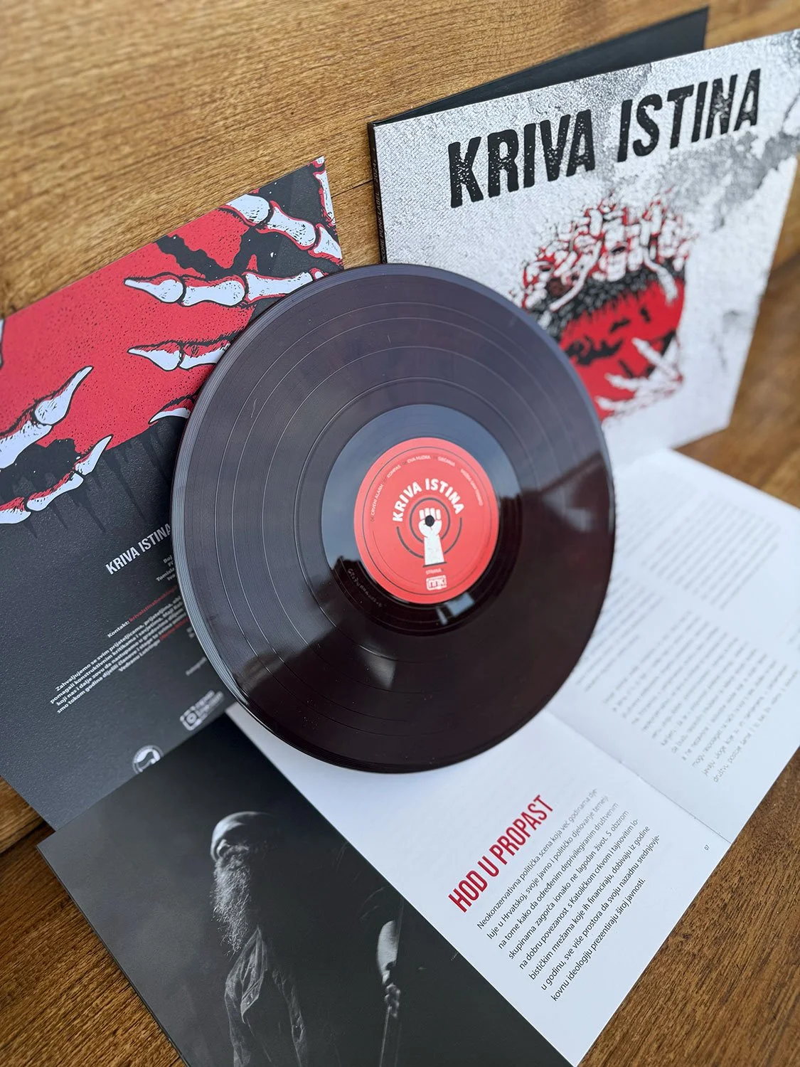 A black vinyl record titled 'KRIVA ISTINA' is placed on an open booklet, with its cover and a page with text visible. The record's center label has a red background with a white raised fist symbol. 