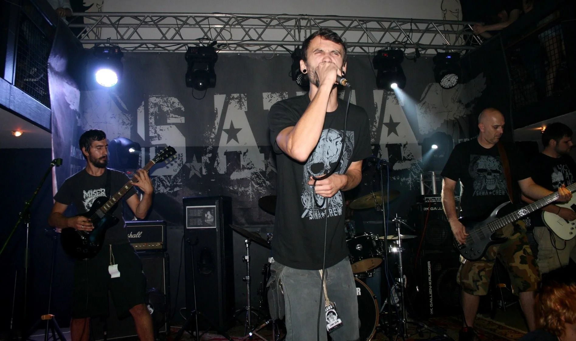 A band performing on stage, with the lead singer in the center holding a microphone and wearing a black T-shirt with a graphic design. To the left, a guitarist with a dark guitar and a beard, and to the right, two other band members playing guitars, 