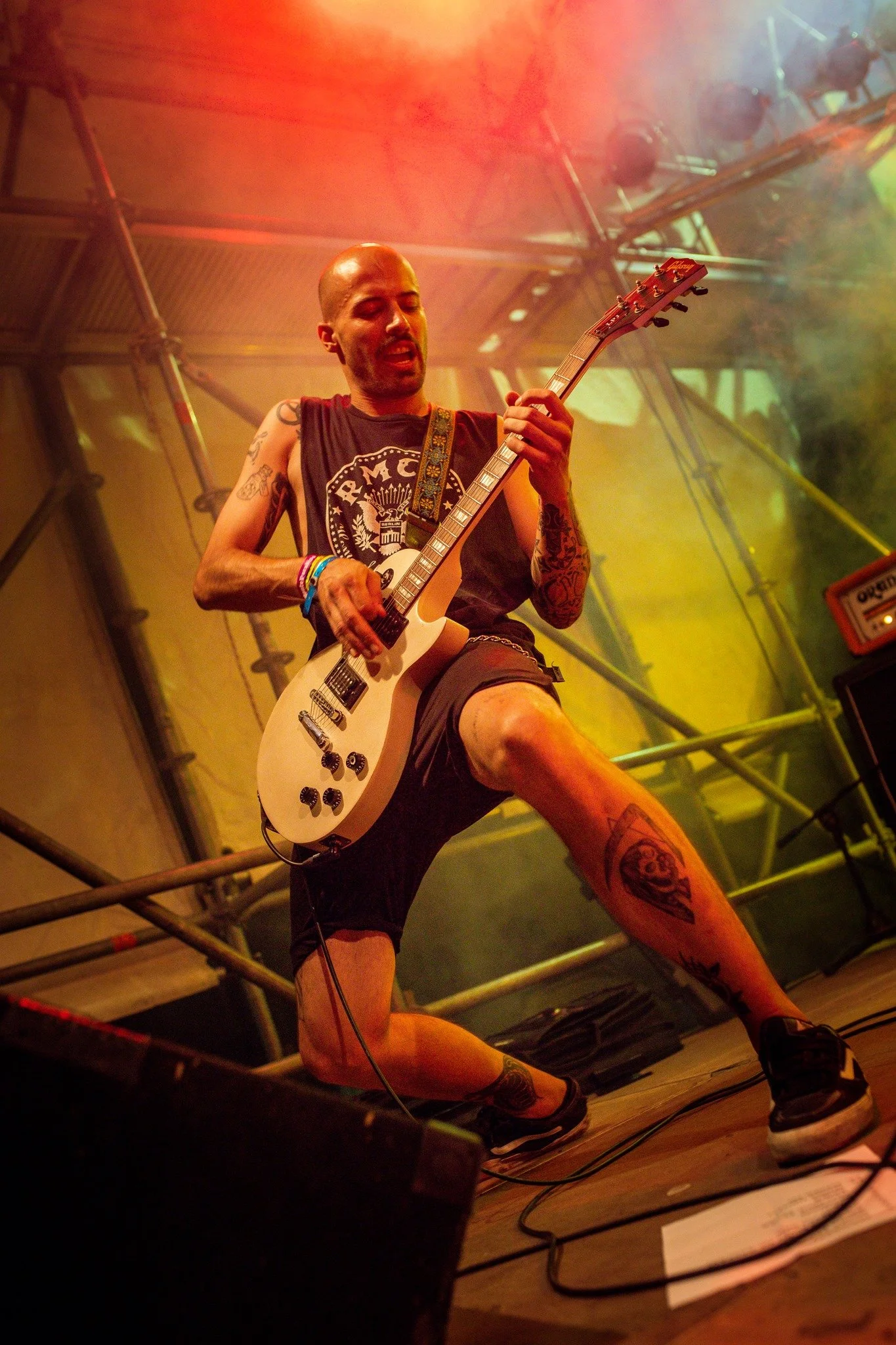 Musician playing an electric guitar on stage under colorful stage lighting with visible scaffolding.
