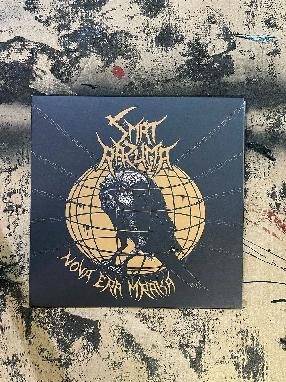 Album cover with a black background, a stylized raven sketch in front of a large yellow globe, chains extending from the globe, and gold text reading "Smrt Razuma" and "Nova Era Mraka".