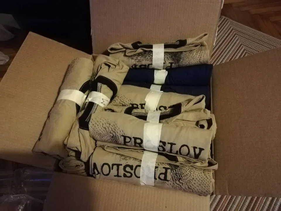 Open cardboard box containing folded beige T-shirts with black text and design, some with navy blue T-shirts, secured with white plastic straps.