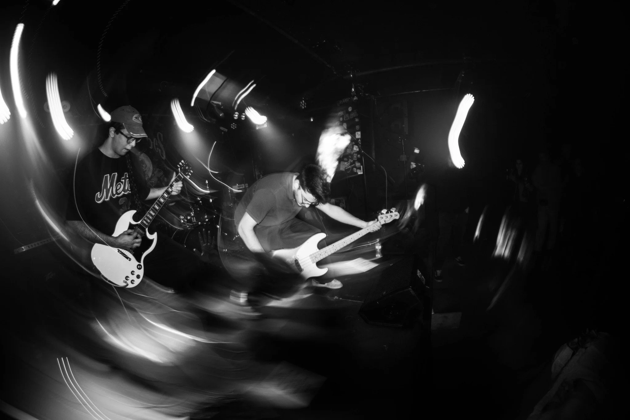 Black and white photo of two musicians playing guitars on stage, with light trails creating a sense of movement and a dark background.