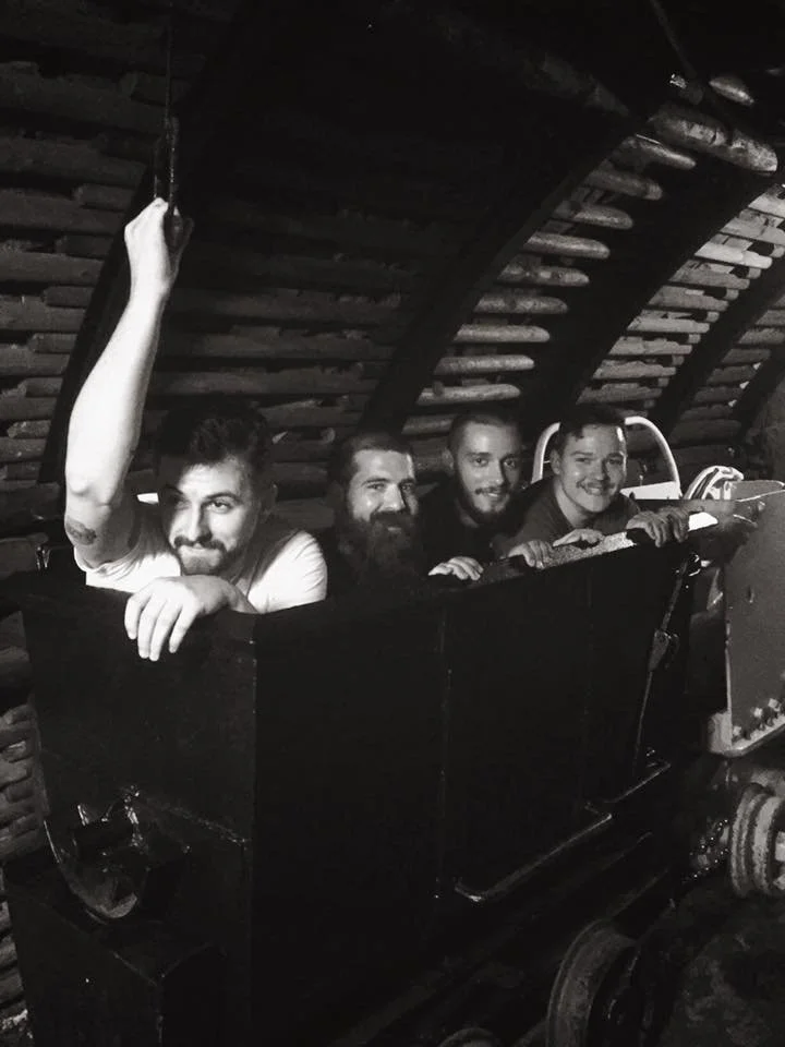 Four people riding in a roller coaster car inside a wooden structure, with one person raising their arm.