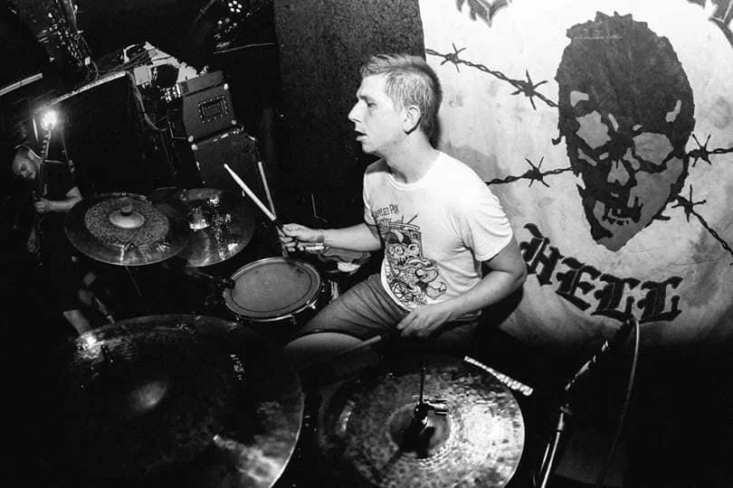 A young man playing a drum set in a dark room with a banner displaying a skull graphic and barbed wire.