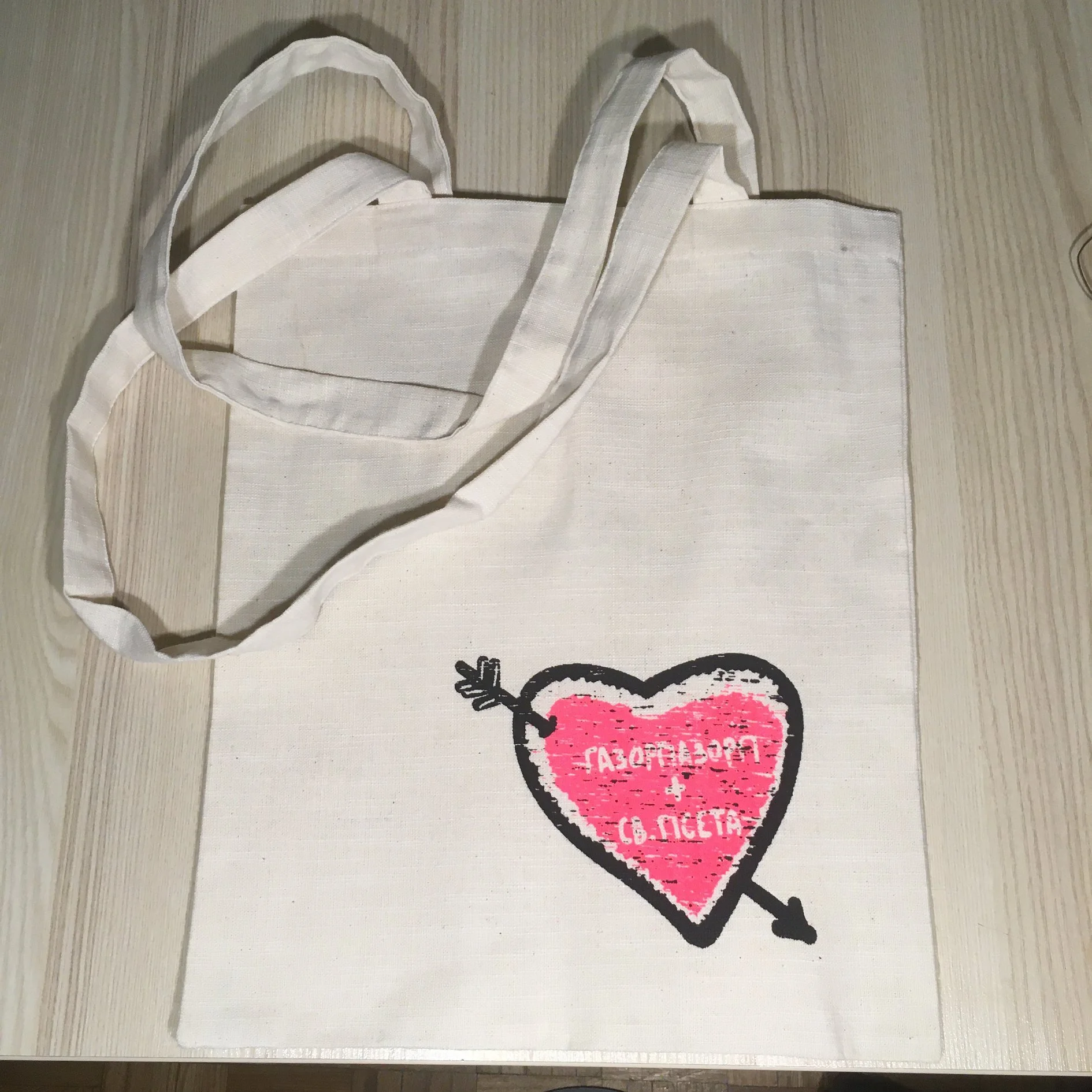 A white fabric tote bag lying flat on a light-colored wooden surface, with handles extended upward. Part of a pink and black heart-shaped graphic with text is visible beneath the bag.