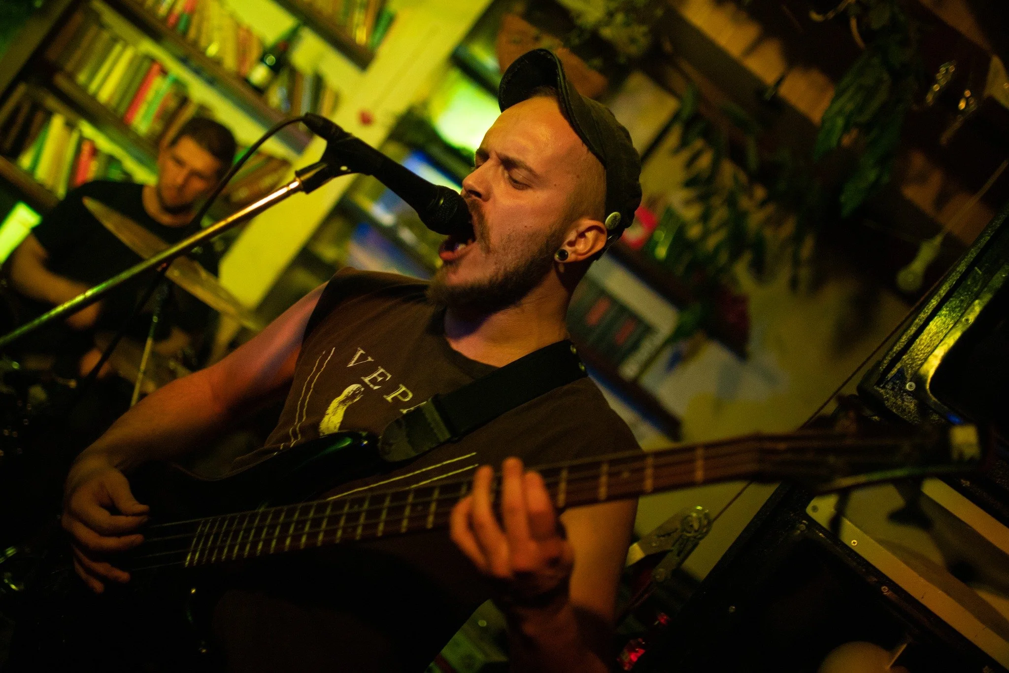 A man singing into a microphone while playing an acoustic guitar in a dimly lit room, with another person in the background.