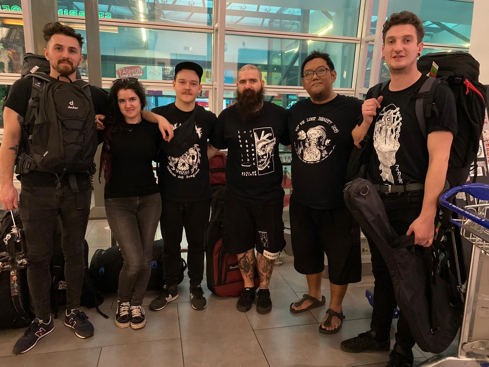 A group of six diverse young adults standing together at an airport, posing for a photo with all carrying backpacks and luggage.