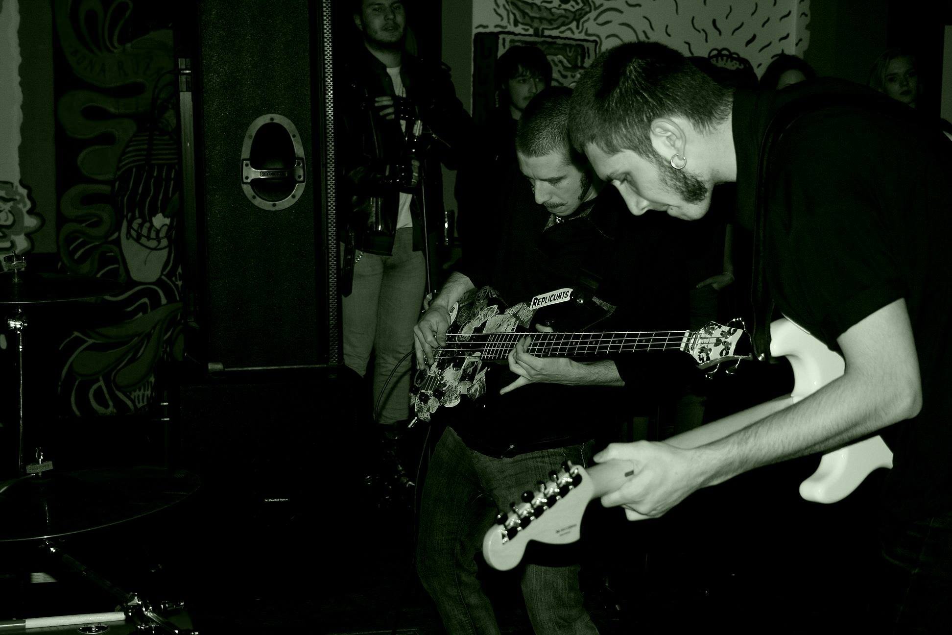 People in a dimly lit room gathered around a person sitting and playing an electric guitar with stickers. Other individuals stand around watching, with one person holding a beer bottle. The room has dark, artistic wall decor.
