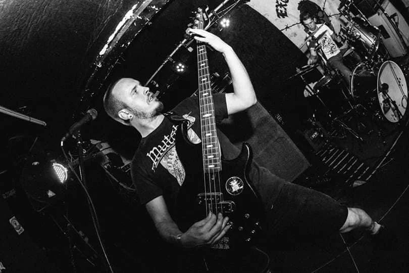 A musician playing an electric bass guitar on stage during a live concert, with another musician playing drums in the background.