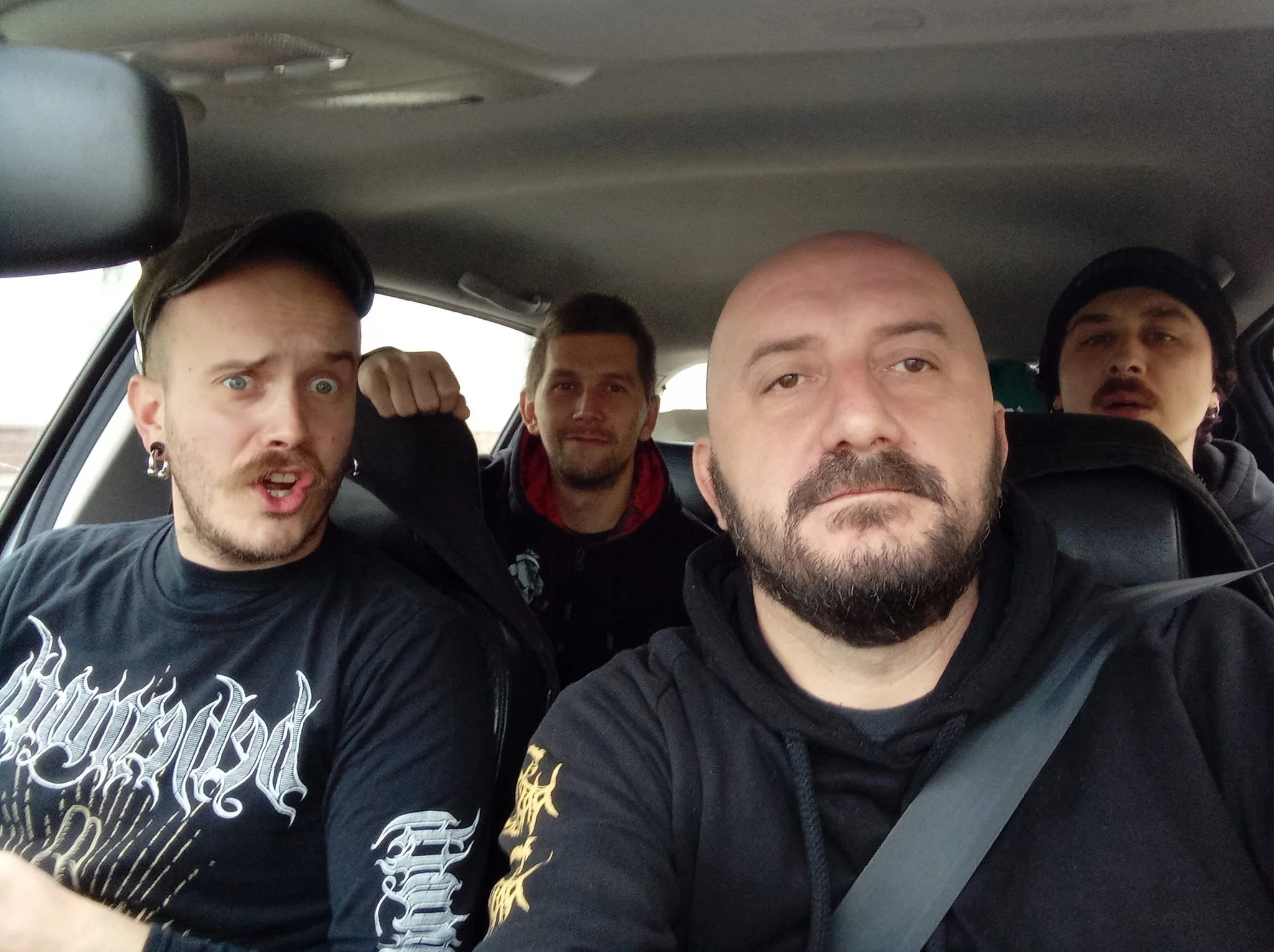 Four men sitting in a car, taking a selfie. The man in the front right has a beard and is missing hair. The man in front left is wearing earrings and a cap. The two men in the back are partially visible, one with short hair and the other wearing a ha