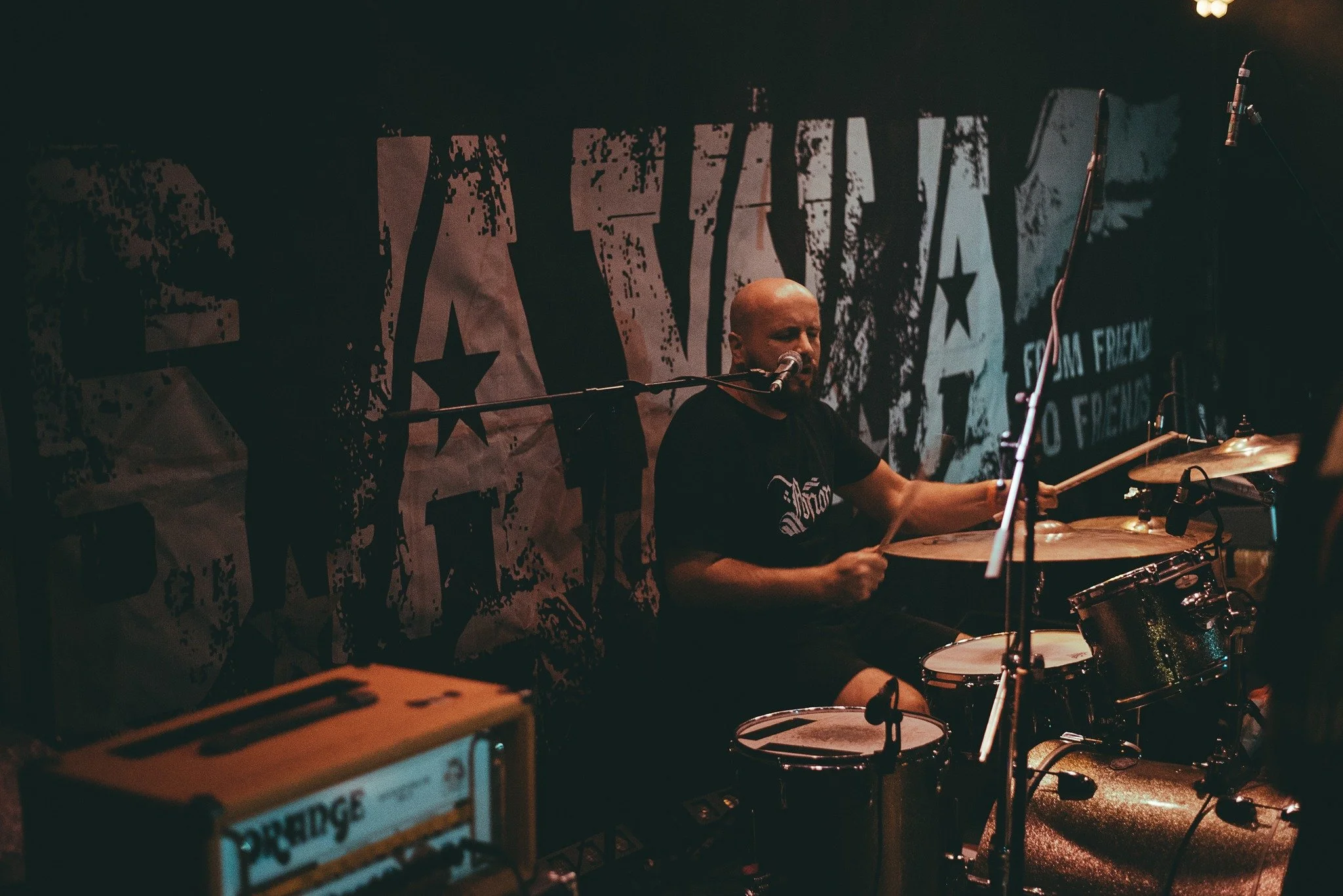 A person playing drums on stage at a music venue with a large black and white graffiti-style backdrop featuring stars and text. The musician has a shaved head and is singing into a microphone while playing the drums.