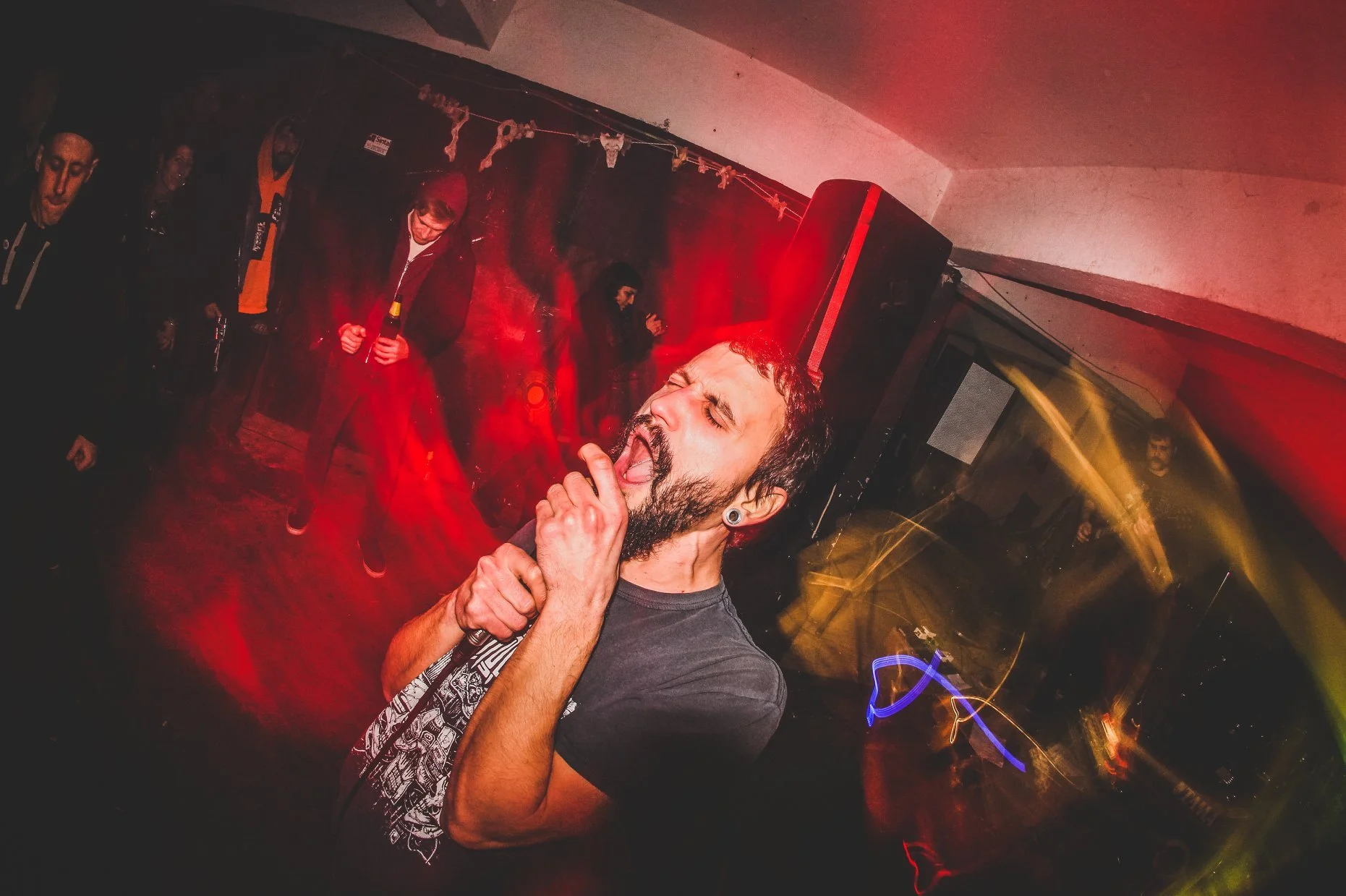 A man with a beard and earrings singing passionately into a microphone at a concert, with his eyes closed and hand near his face. In the background, a group of people are standing against a red-lit wall, some with hands clasped or holding drinks, in 