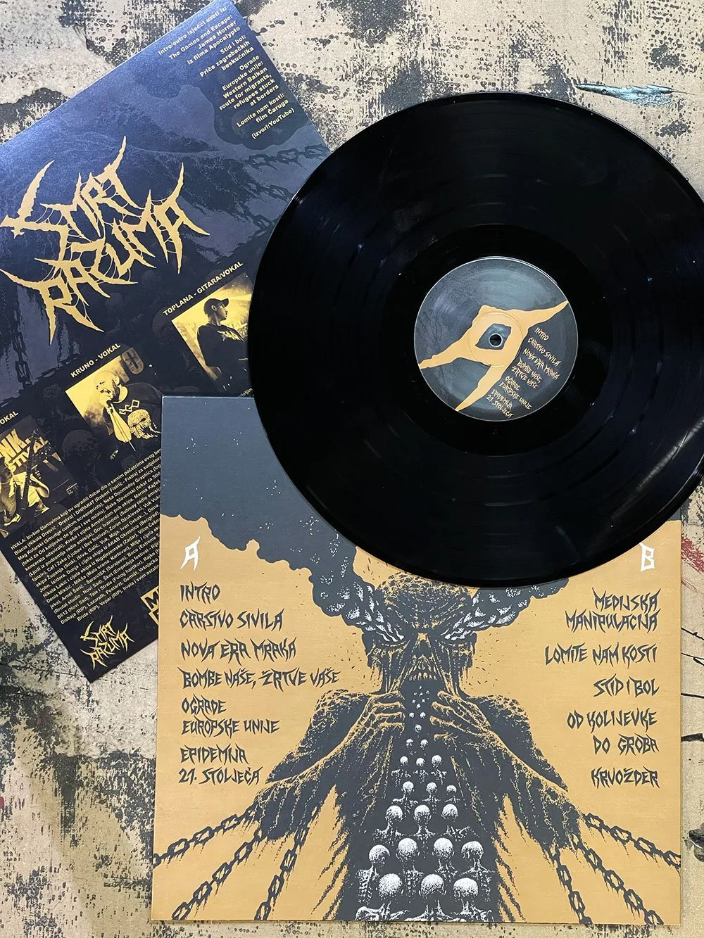 A black vinyl record resting on a poster featuring a graphic of a mushroom cloud with skulls at its base. The poster has handwritten-style text listing song titles and band members, with some additional printed information and a small photo insert. T