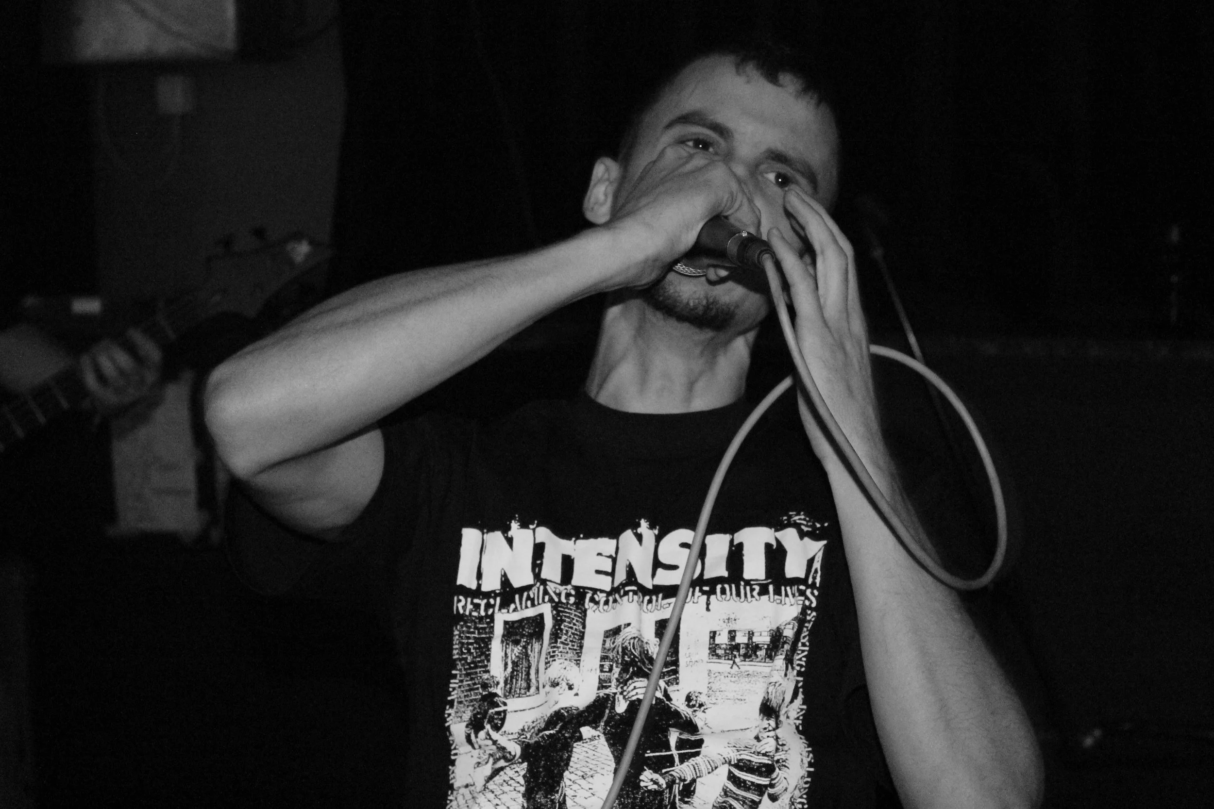 A young man with short dark hair is screaming at a dark indoor venue. He is wearing a black T-shirt with white text and a graphic design. There is a person next to him holding a guitar, but only their arm and part of the guitar are visible. 