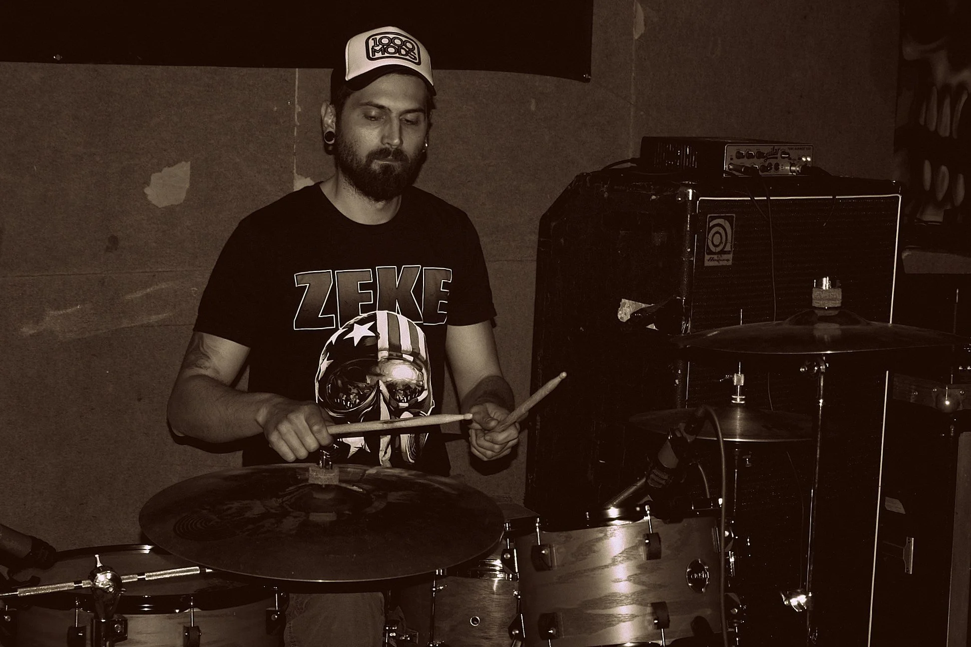 A man playing the drums in a music studio, wearing a black T-shirt with a skull design and a white baseball cap with the logo '1000 Mods', surrounded by musical equipment.