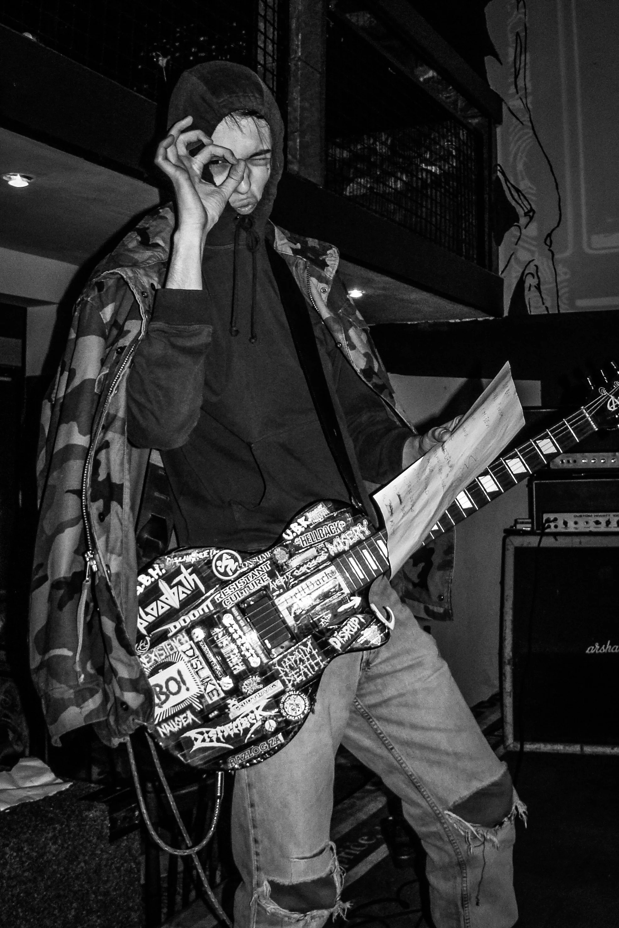 Person wearing hoodie and camouflage jacket holding a guitar decorated with stickers, making a playful face and putting their hand around one eye in a circle gesture, in a music venue with amplifiers and a railing in the background.