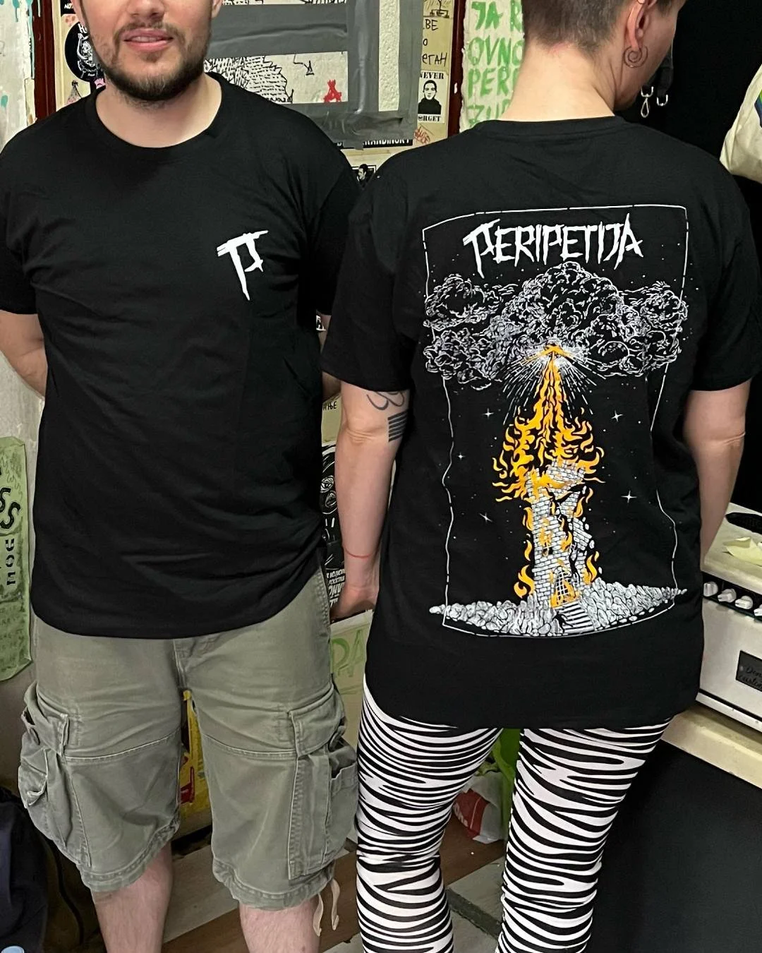 Two people standing side by side. The person on the left is wearing a black T-shirt with a small white logo. The person on the right is wearing a black T-shirt with a large graphic design on the back, featuring a smoky explosion.