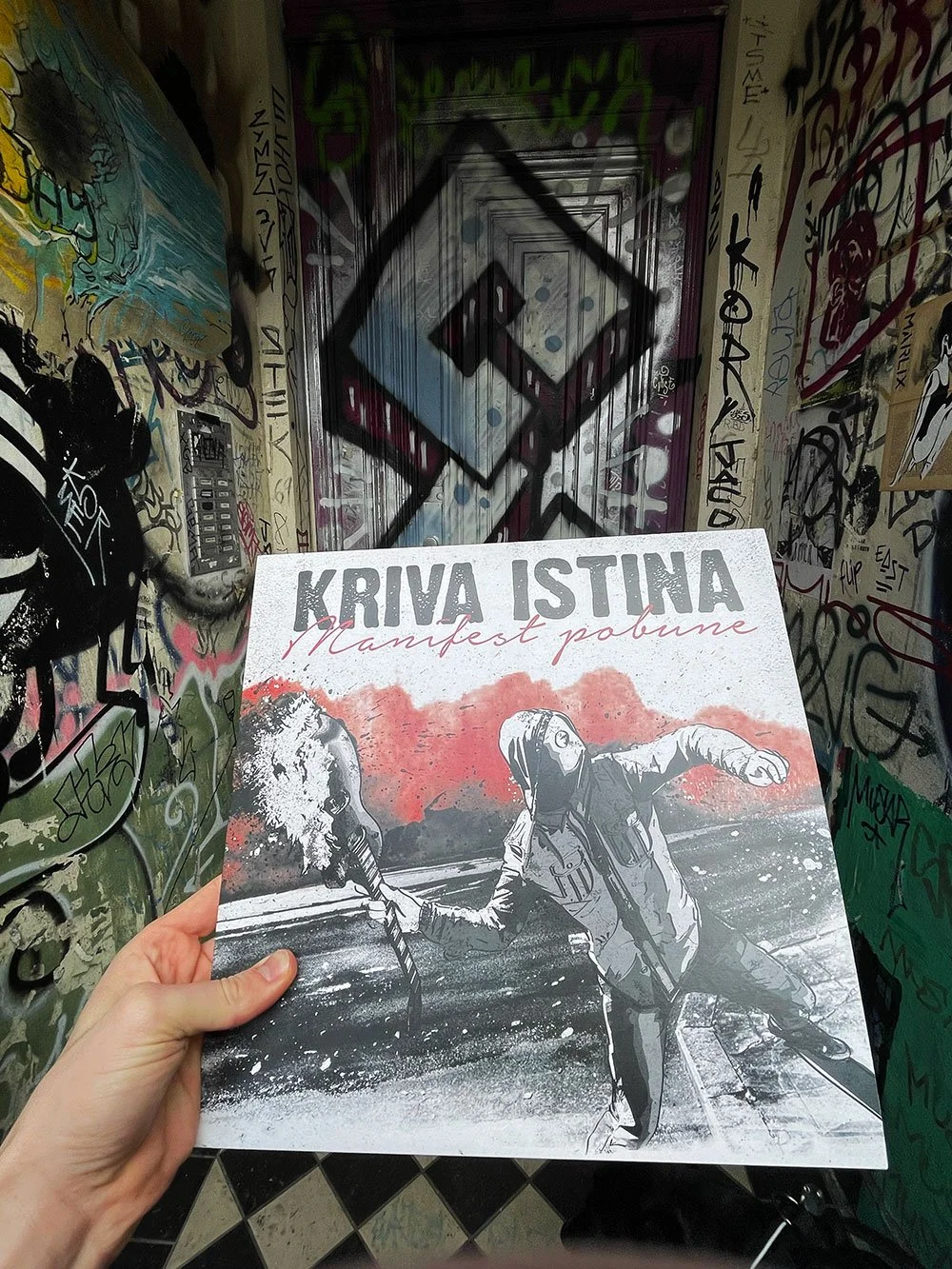 A person holding a protest flyer with animated style art of a woman wearing a gas mask, holding a flaming torch, in a graffiti-covered stairwell. The flyer has the text 'KRIVA ISTINA' and 'Manifest pobune'.
