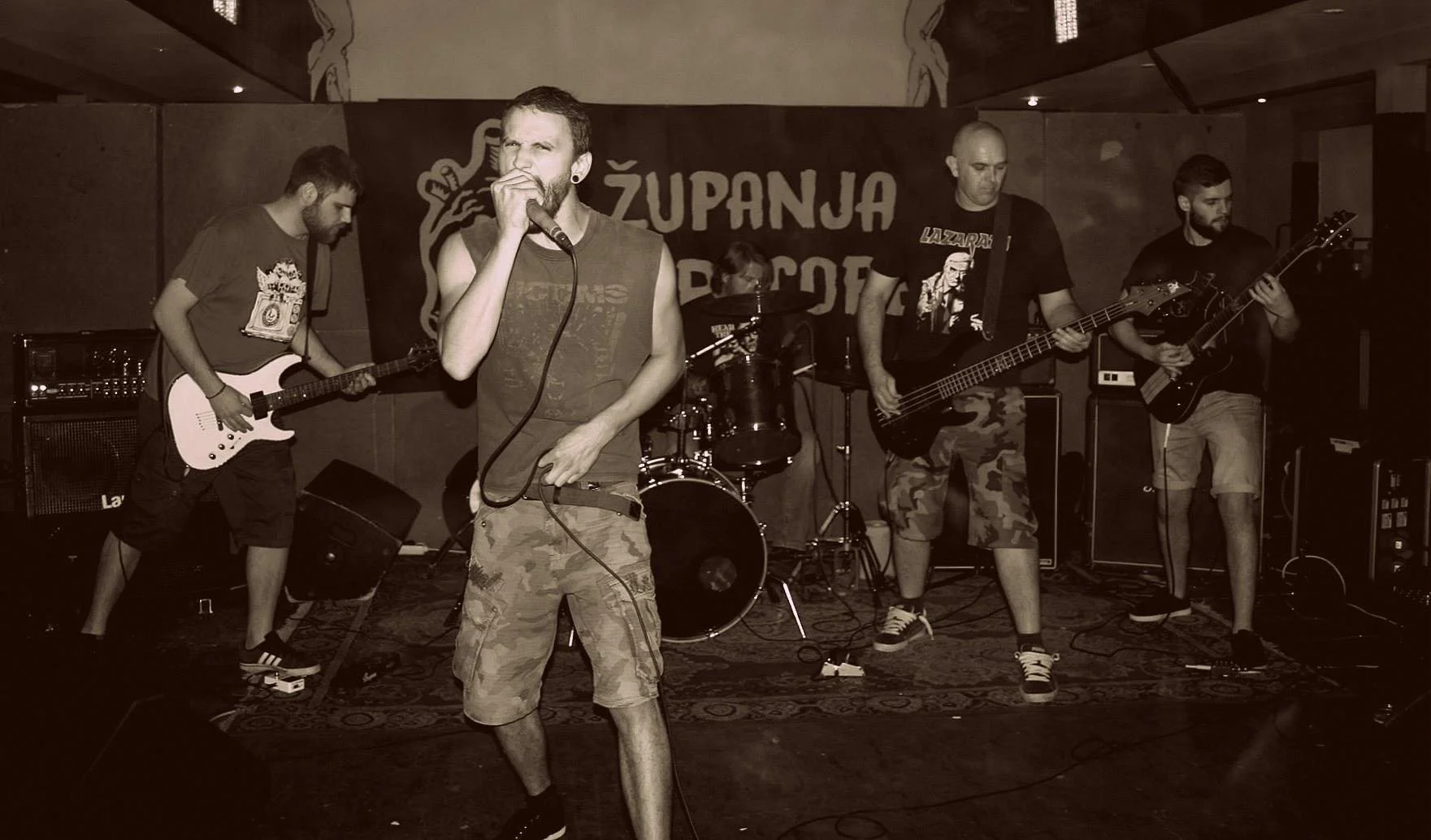 A band performing live on stage with five members: a vocalist holding a microphone, two guitarists, a bassist, and a drummer in the background. The band members are dressed casually in t-shirts and shorts. There is a drum set, amplifiers, and a backd