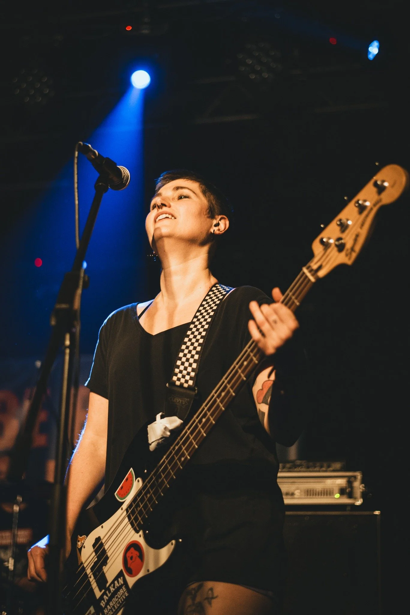 A woman with short hair singing into a microphone while playing an electric bass guitar on stage, with blue stage lights overhead and a music amplifier in the background.