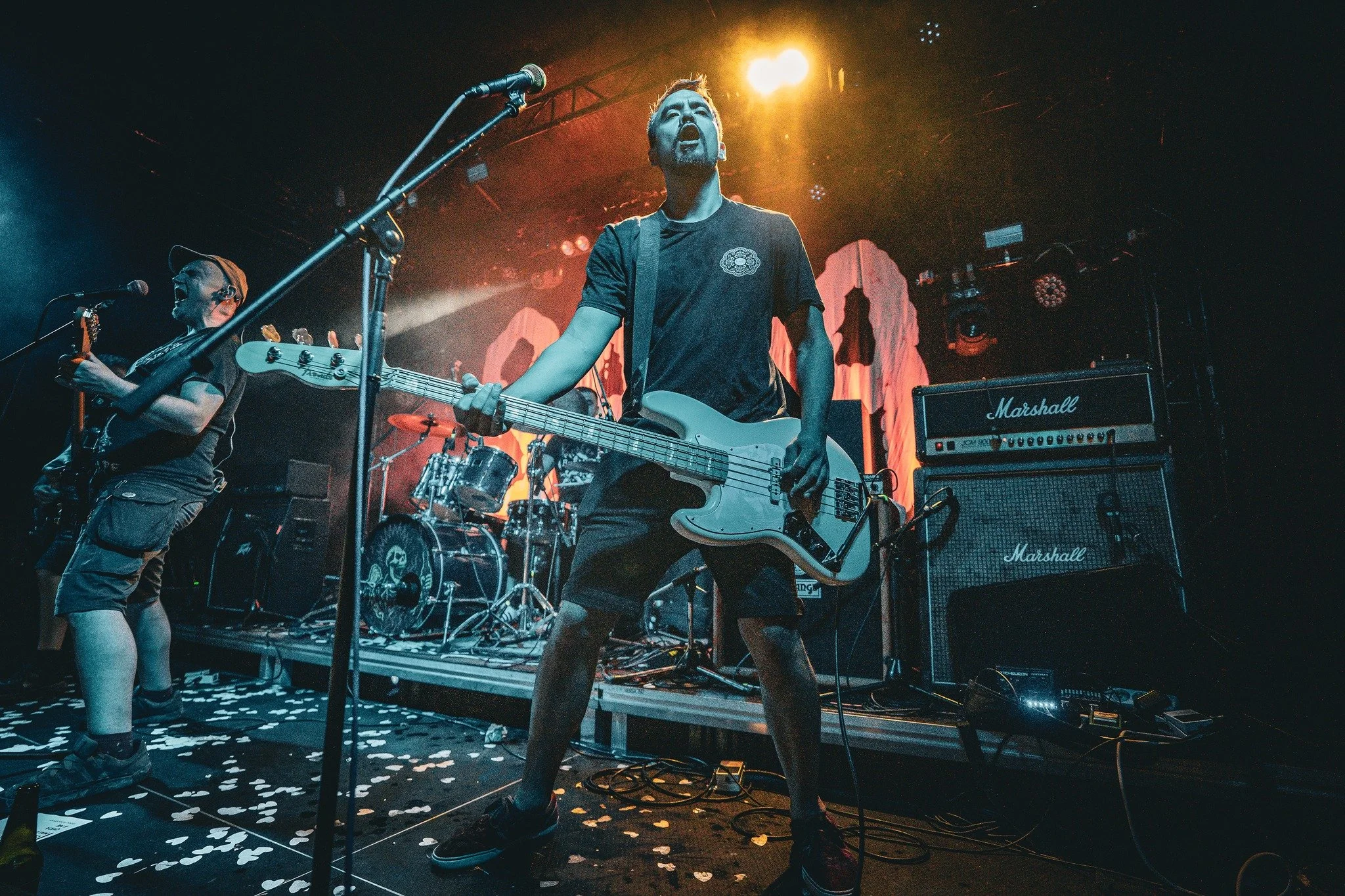 Musicians performing on stage at a concert with bright stage lights, including a guitarist and bassist, with equipment like Marshall amplifiers, and confetti on the floor.
