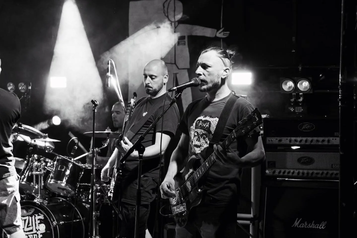 Black and white photo of a live band performance featuring three male musicians on stage, with a drummer at the back, and the lead singer and guitarist in the front, musical equipment, overhead lights, and a dark background.