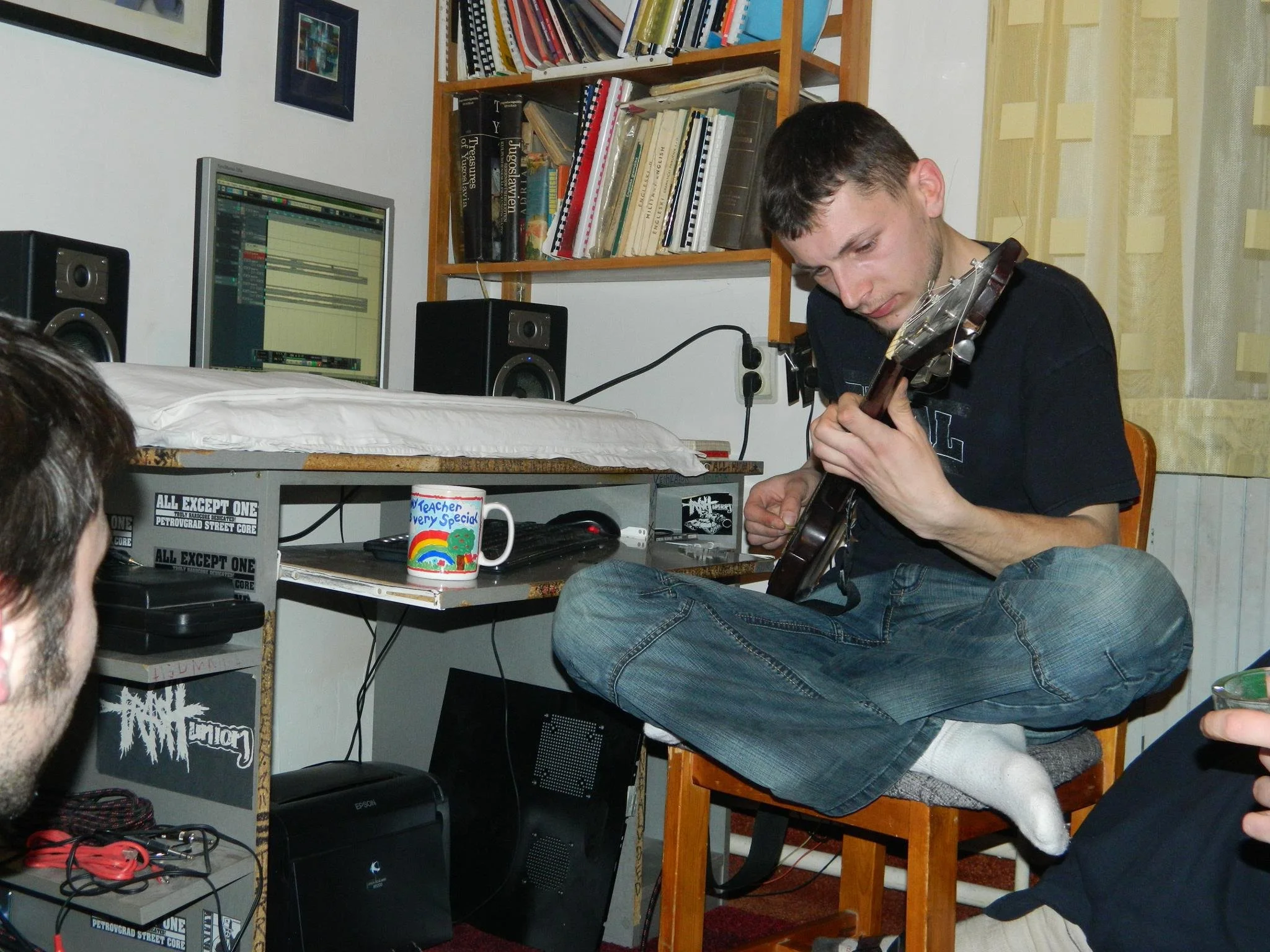 Young man playing guitar while sitting on a wooden table in a room with bookshelves, computer, and colorful mug.