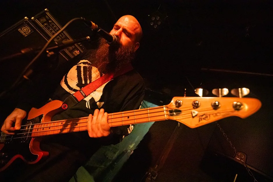 A bearded man singing into a microphone while playing an electric bass guitar on stage.