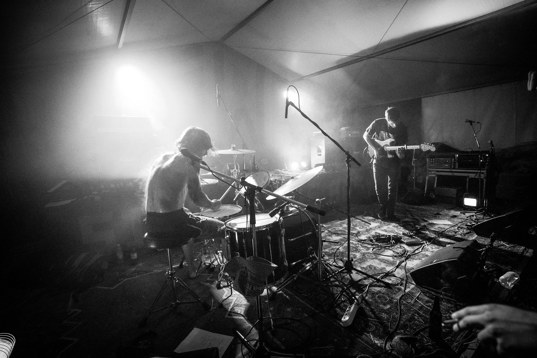 A black-and-white photo of a live music performance featuring two musicians playing instruments on stage, with bright stage lights behind them creating a hazy atmosphere.