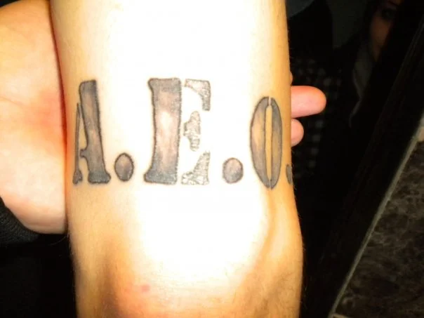 Tattoo on person's arm with the text AEO in bold, black lettering.