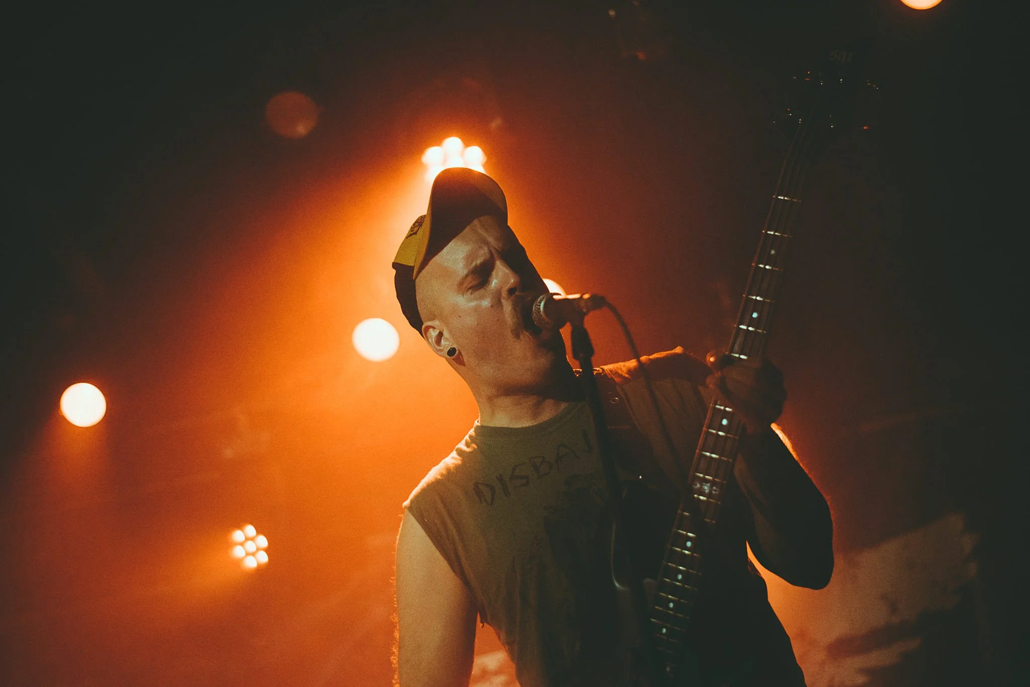 A musician with a cap and earrings singing into a microphone while playing an electric guitar on stage at night with orange stage lighting.