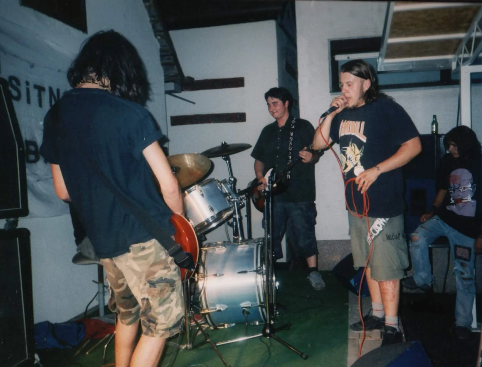 A band of young musicians performing on stage with drums and guitars, in a casual indoor setting.