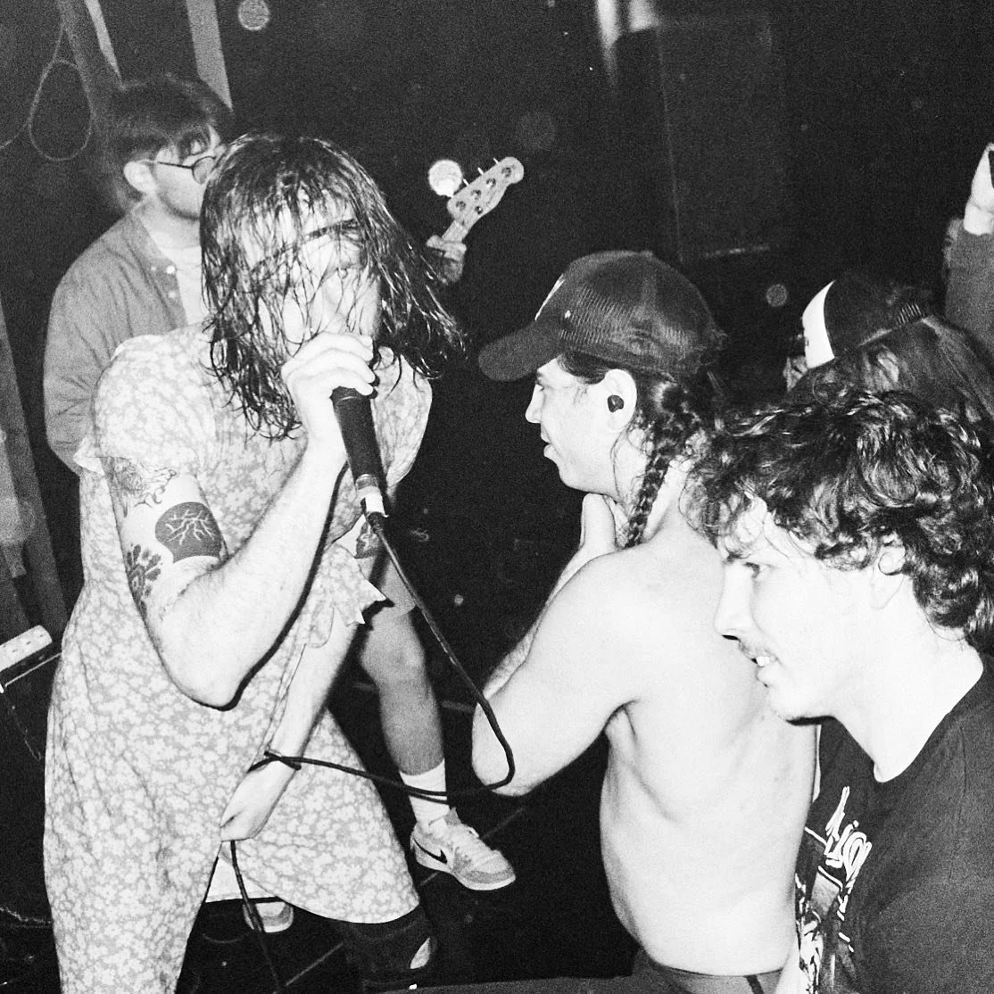 A black and white photo of a punk rock band performing on stage, with a singer with long, wet hair holding a microphone, a shirtless guitarist with a cap, and two other band members with curly hair wearing glasses, all intensely engaged in the perfor