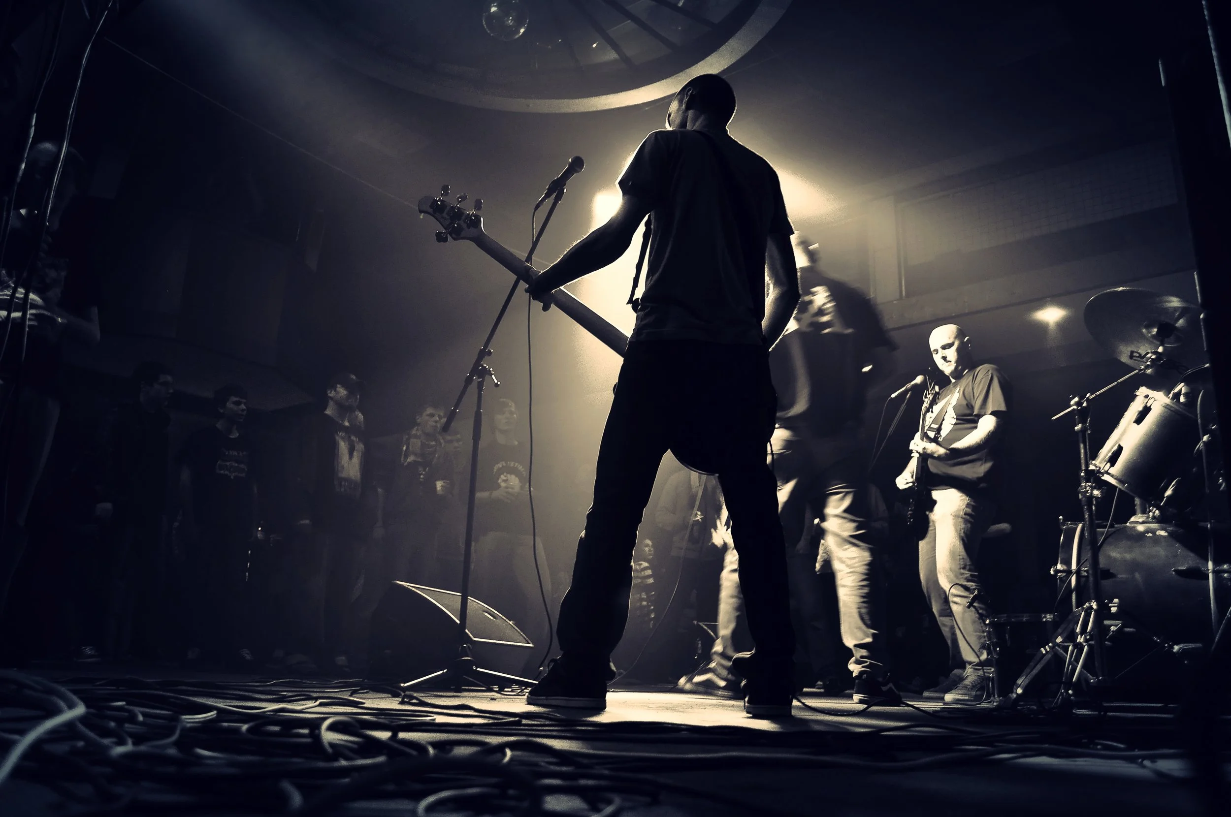 A musician playing guitar on stage with an audience in front, in a dark, moody setting illuminated by backlighting.